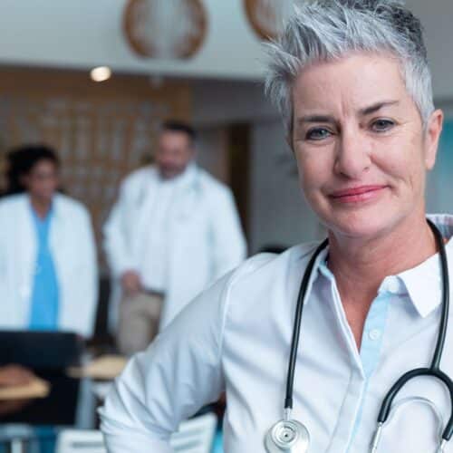 Older female caucasian doctor with stethoscope in foreground, other medical professionals blurry in background