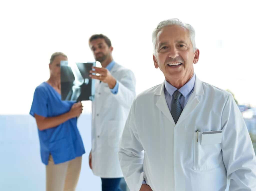 Mature caucasian doctor smiling in foreground, doctors looking at an x-ray in background